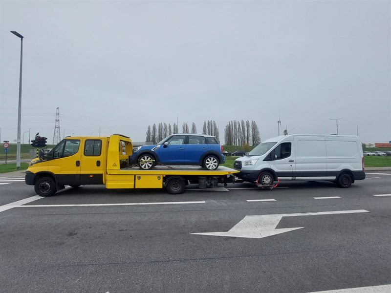 Yellow flatbed truck carrying a blue Mini and towing a white van